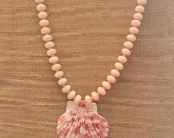 Può includere: Una collana composta da una fila di perline rotonde rosa pallido infilate su un cordino rosa, con al centro un grande ciondolo a forma di conchiglia a ventaglio. La conchiglia presenta un motivo naturale di strisce radiali bianche e rosa.
