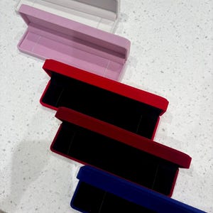May include: A selection of open jewellery boxes in different colours. The boxes are rectangular with a hinged lid and a soft, velvet interior. Colours include white, pink, red, and blue. The boxes are designed to hold necklaces or bracelets.