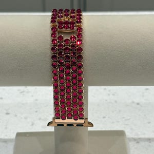 May include: A rose gold Apple Watch strap embellished with multiple rows of bright red gemstones. The strap is displayed on a neutral stand, highlighting its sparkling design and elegant aesthetic.