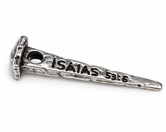 May include: A silver-toned metal nail pendant with a textured, rustic finish. The head of the nail is flat and square-shaped, and the shaft features a circular hole near the top. The text ISAIAS 53:6 is embossed along the length of the nail shaft.