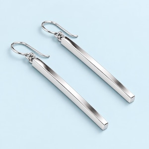 May include: A pair of silver-toned drop earrings featuring long, slender, rectangular bar pendants suspended from simple curved ear wires. The metal surfaces are smooth and reflective, catching the light to highlight the clean, straight edges of the bars.