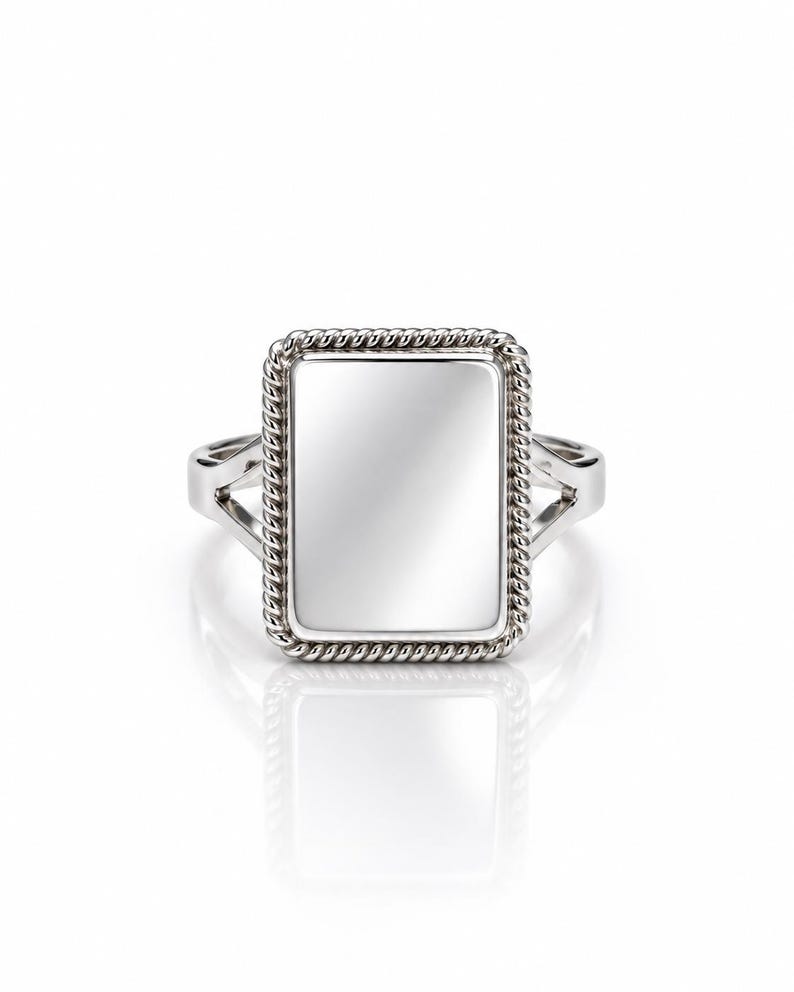 May include: A silver-toned ring featuring a rectangular face with a smooth, reflective center panel. The rectangular centerpiece is framed by a textured, twisted rope-style border. The band of the ring splits into two sections.