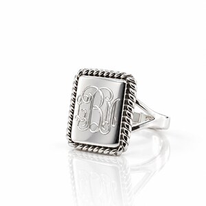 May include: A silver-toned ring featuring a rectangular face with a polished surface. The center of the face is engraved with the script monogram letters BM. The rectangular face is framed by a twisted rope-style border and a split band.