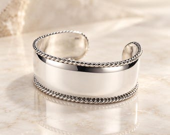 May include: A polished silver cuff bracelet featuring a wide, smooth central band bordered by two twisted rope-style edges. The open-ended design is crafted from a reflective metallic material, showcasing a clean, minimalist aesthetic.