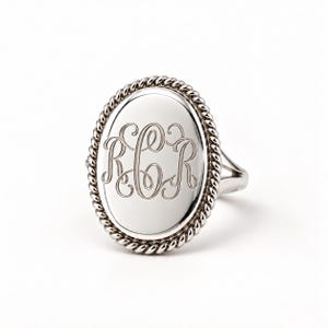 May include: A silver-toned ring featuring an oval face with a twisted rope-style border. The center of the oval is smooth and polished, engraved with the ornate, interconnected script letters R, C, and R in a classic monogram style.