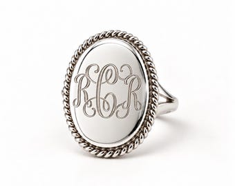 May include: A silver-toned ring featuring an oval face with a twisted rope-style border. The center of the oval is smooth and polished, engraved with the ornate, interconnected script letters R, C, and R in a classic monogram style.