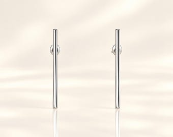 May include: A pair of silver-toned bar earrings featuring a long, slender, vertical cylindrical design. Each earring has a smooth, polished metallic finish and includes a small circular backing post attached to the upper portion of the bar.