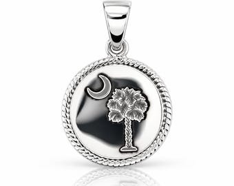 May include: A circular silver pendant featuring a polished finish with a raised rope-style border. The center displays an engraved design of a crescent moon and a palmetto tree. The pendant includes a tapered silver bail at the top for attachment.