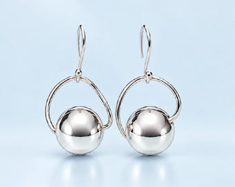 May include: A pair of silver-toned drop earrings featuring a polished, spherical bead suspended within a curved, open circular frame. The earrings are attached to a simple hook-style finding, showcasing a smooth, reflective metallic finish throughout.