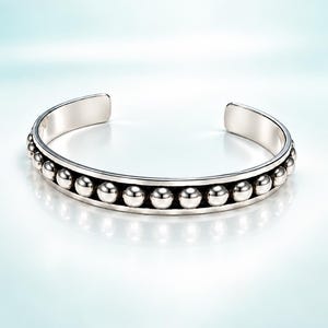 May include: A silver-toned cuff bracelet featuring a central row of polished spherical beads set within a smooth, recessed channel. The open-ended design has a reflective metallic finish and a consistent, rounded band structure throughout the piece.