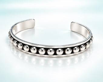 May include: A silver-toned cuff bracelet featuring a central row of polished spherical beads set within a smooth, recessed channel. The open-ended design has a reflective metallic finish and a consistent, rounded band structure throughout the piece.