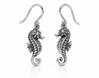 May include: A pair of silver-toned seahorse earrings featuring intricate textured detailing on the bodies, fins, and curled tails. Each seahorse is suspended from a curved hook wire, showcasing a polished metallic finish with darkened recessed areas.