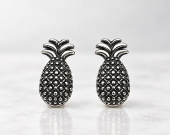May include: A pair of silver-toned stud earrings shaped like pineapples. Each earring features a textured, dotted surface on the fruit body and smooth, layered leaves at the top, finished with a polished metallic sheen.