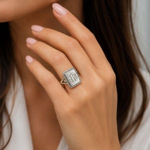May include: A silver-toned rectangular signet ring featuring a twisted rope-style border and an engraved monogram with the letters BM in the center. The ring has a split shank design and a polished metallic finish on the face.