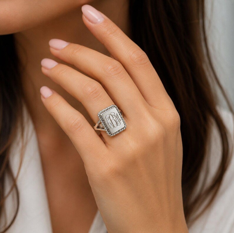 May include: A silver-toned rectangular signet ring featuring a twisted rope-style border and an engraved monogram with the letters BM in the center. The ring has a split shank design and a polished metallic finish on the face.