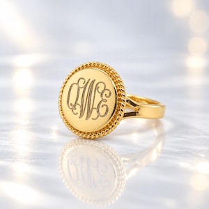 May include: A gold-toned ring featuring a circular face with a rope-textured border. The center of the disc is engraved with the stylized cursive letters QWE in a classic monogram style. The band is smooth and connects to the circular setting.
