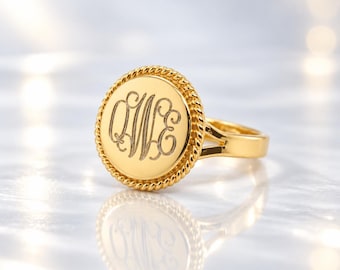 May include: A gold-toned ring featuring a circular face with a rope-textured border. The centre of the disc is engraved with the stylised cursive letters QWE in a classic monogram style. The band is smooth and connects to the circular setting.