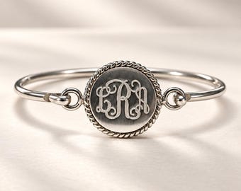 May include: A silver-toned bangle bracelet featuring a central circular charm with a rope-style border. The charm is engraved with the interlocking script letters LRA. The bracelet has a smooth, polished finish and a hinged connection to the charm.