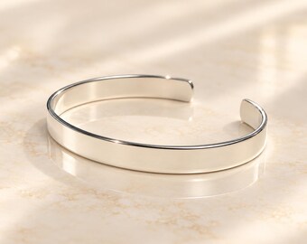 May include: A polished silver-tone cuff bracelet with a smooth, flat surface and an open-ended design. The bracelet features a reflective, metallic finish that catches the light, showcasing a clean, simple, and continuous band structure.