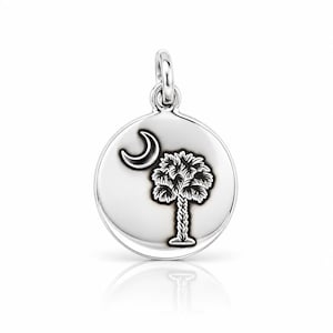 May include: A round silver pendant featuring an engraved design of a crescent moon and a palmetto tree. The charm has a polished, reflective surface with dark, recessed lines highlighting the detailed palm tree and moon imagery.