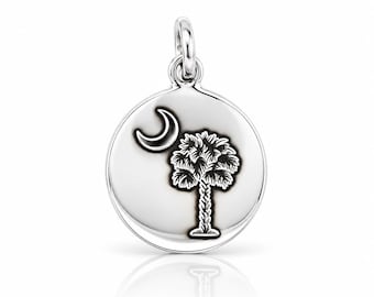 May include: A round silver pendant featuring an engraved design of a crescent moon and a palmetto tree. The charm has a polished, reflective surface with dark, recessed lines highlighting the detailed palm tree and moon imagery.