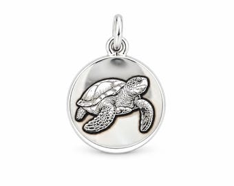 May include: A circular silver pendant featuring a detailed, engraved black and white illustration of a sea turtle swimming. The pendant has a polished metallic finish and includes a small silver jump ring at the top for attachment.
