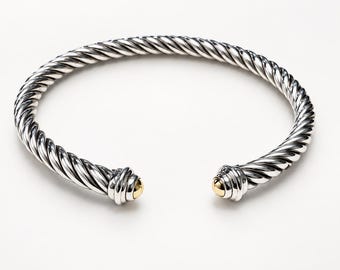 May include: A silver cuff bracelet featuring a twisted cable design throughout the band. The open ends of the bracelet are finished with polished silver caps and smooth, rounded gold-toned accents at the very tips of the metal structure.