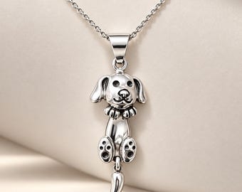 May include: A silver-toned metal pendant necklace featuring a stylized dog figure with floppy ears, a small collar, and visible paw pads on its feet. The dog is depicted in a vertical hanging position, suspended from a delicate silver-toned chain.