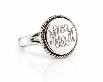 May include: A silver-toned ring featuring a circular face with a rope-style border. The center of the disc is engraved with the monogram letters MCM in a cursive script. The band of the ring has a split design where it meets the circular face.