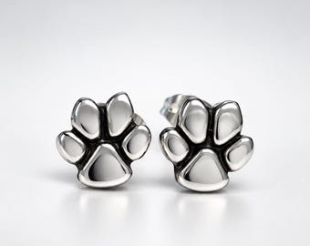May include: A pair of silver-toned stud earrings shaped like animal paw prints. Each earring features a central triangular pad and four rounded toe pads with recessed dark detailing between the segments, creating a polished metallic finish.