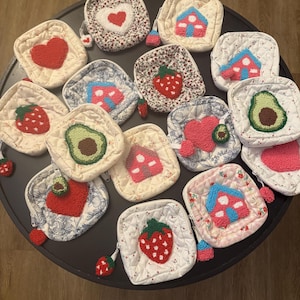 May include: A collection of quilted, square pouches with various embroidered designs, including hearts, houses, strawberries, and avocados. The pouches are white with floral or patterned backgrounds, and some have small pom-pom zipper pulls. The color palette includes red, pink, green, and blue.