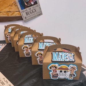May include: A row of brown cardboard gable boxes, each decorated with a cutout of the anime character Monkey D. Luffy, a skull and crossbones emblem, and the name MATEO in blue letters. The boxes are arranged on a black surface with posters.