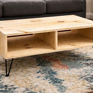 May include: A light-coloured wooden coffee table with a rectangular design. The table has two open storage compartments and sits on black hairpin legs. The table is approximately 122 cm long and 46 cm high.