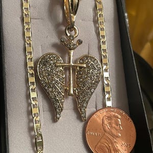 May include: Gold-coloured necklace with a pendant featuring a cross and wing design, encrusted with clear rhinestones. The chain has a flat, linked design. A penny is included for size reference.