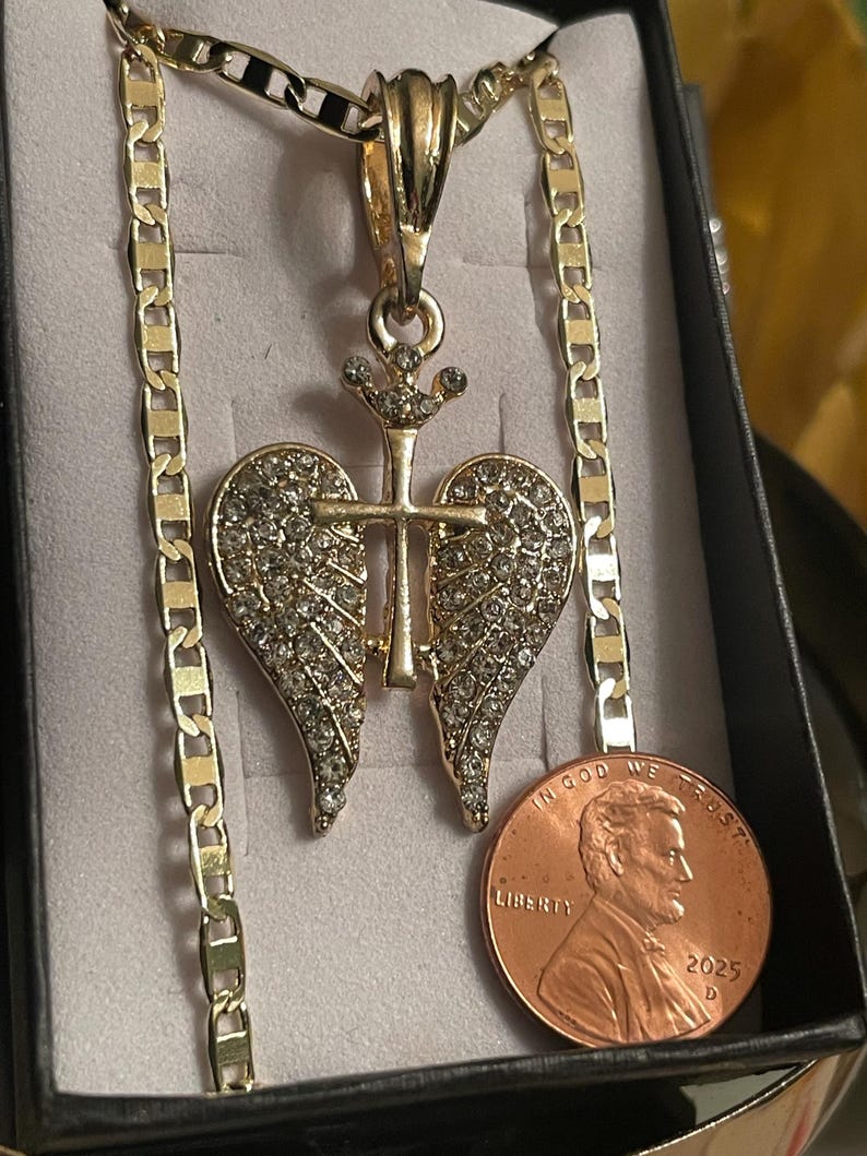 May include: Gold-coloured necklace with a pendant featuring a cross and wing design, encrusted with clear rhinestones. The chain has a flat, linked design. A penny is included for size reference.