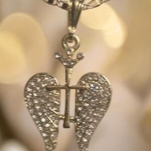 May include: A gold-coloured pendant necklace. The pendant features a cross design with angel wings, both embellished with small crystals. The necklace hangs from a fine chain, offering a classic and elegant appearance.