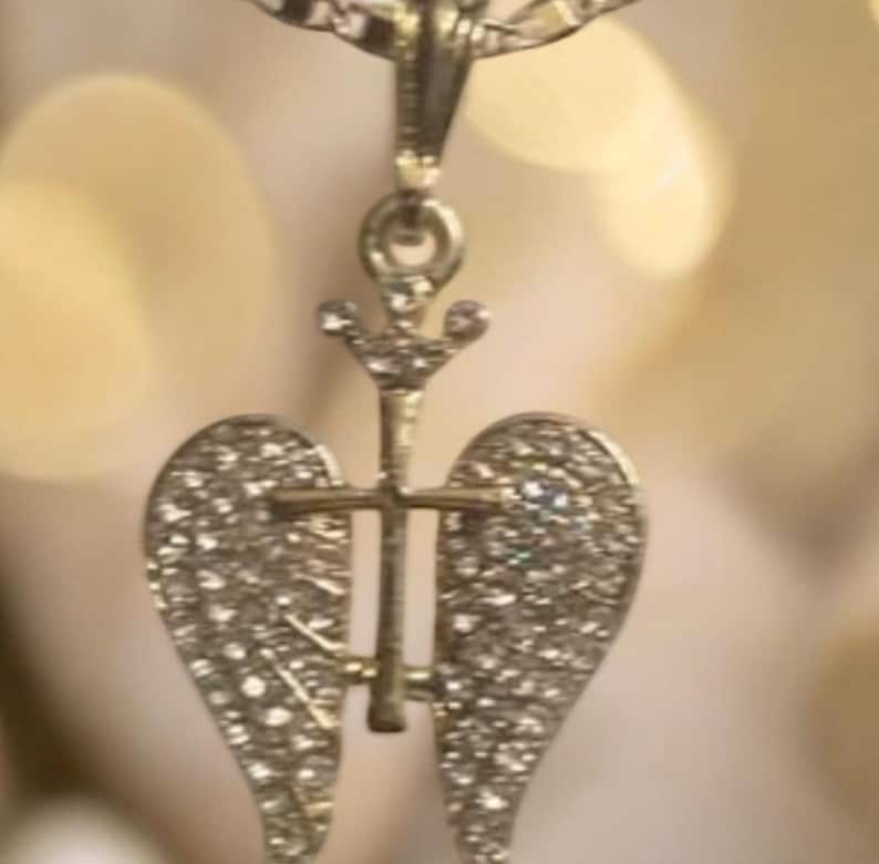 May include: A gold-coloured pendant necklace. The pendant features a cross design with angel wings, both embellished with small crystals. The necklace hangs from a fine chain, offering a classic and elegant appearance.