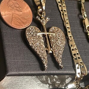 May include: A gold-toned pendant with a cross design, featuring crystal-like embellishments on the wings. A penny is next to the pendant for size comparison. A gold chain is also visible.