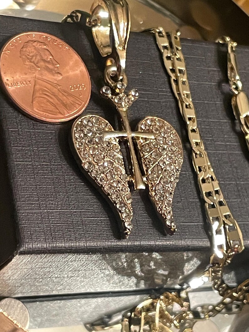 May include: A gold-toned pendant with a cross design, featuring crystal-like embellishments on the wings. A penny is next to the pendant for size comparison. A gold chain is also visible.