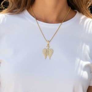 May include: A gold necklace with a pendant shaped like angel wings, set with small, sparkling stones. The pendant has a cross in the middle. The necklace is worn over a white t-shirt.