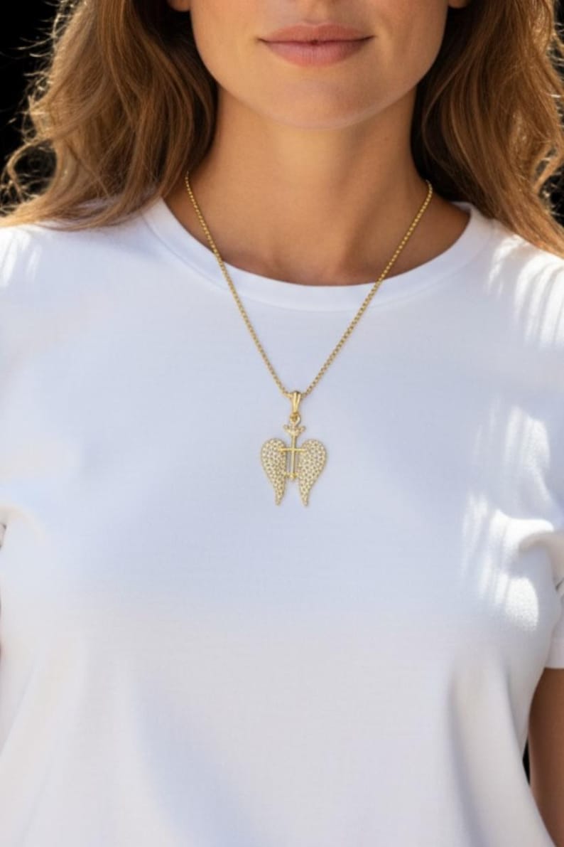May include: A gold necklace with a pendant shaped like angel wings, set with small, sparkling stones. The pendant has a cross in the middle. The necklace is worn over a white t-shirt.