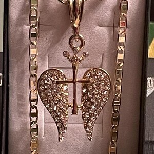 May include: A gold-coloured necklace with a pendant featuring a cross and heart-shaped wings, encrusted with sparkling crystals. The chain is a thick, gold-coloured link design. The pendant is centered on a light-coloured background.