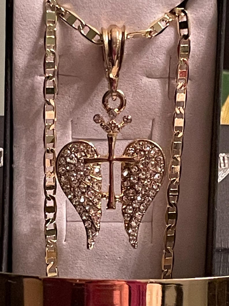 May include: A gold-coloured necklace with a pendant featuring a cross and heart-shaped wings, encrusted with sparkling crystals. The chain is a thick, gold-coloured link design. The pendant is centered on a light-coloured background.