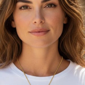 May include: A woman wearing a white t-shirt and a gold necklace with a pendant. The pendant features a cross and wing design, encrusted with small, sparkling stones. The woman has brown hair and eyes.