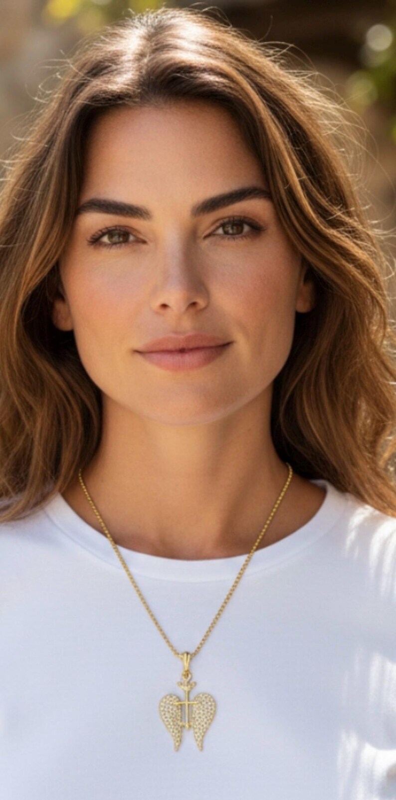 May include: A woman wearing a white t-shirt and a gold necklace with a pendant. The pendant features a cross and wing design, encrusted with small, sparkling stones. The woman has brown hair and eyes.