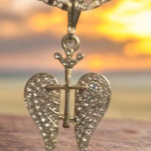 May include: A gold-coloured pendant necklace with an angel wing design. The pendant features a cross and is decorated with small, sparkling stones. The background shows a blurred sunset.