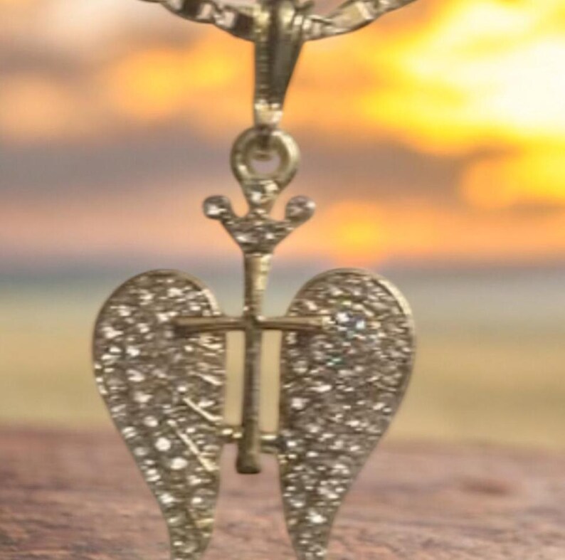 May include: A gold-coloured pendant necklace with an angel wing design. The pendant features a cross and is decorated with small, sparkling stones. The background shows a blurred sunset.