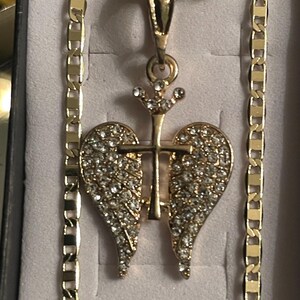 May include: Gold-coloured necklace with a pendant featuring a cross and angel wings design, encrusted with sparkling rhinestones. The chain has a flat, rectangular link pattern. The pendant is centered on a light-coloured background.