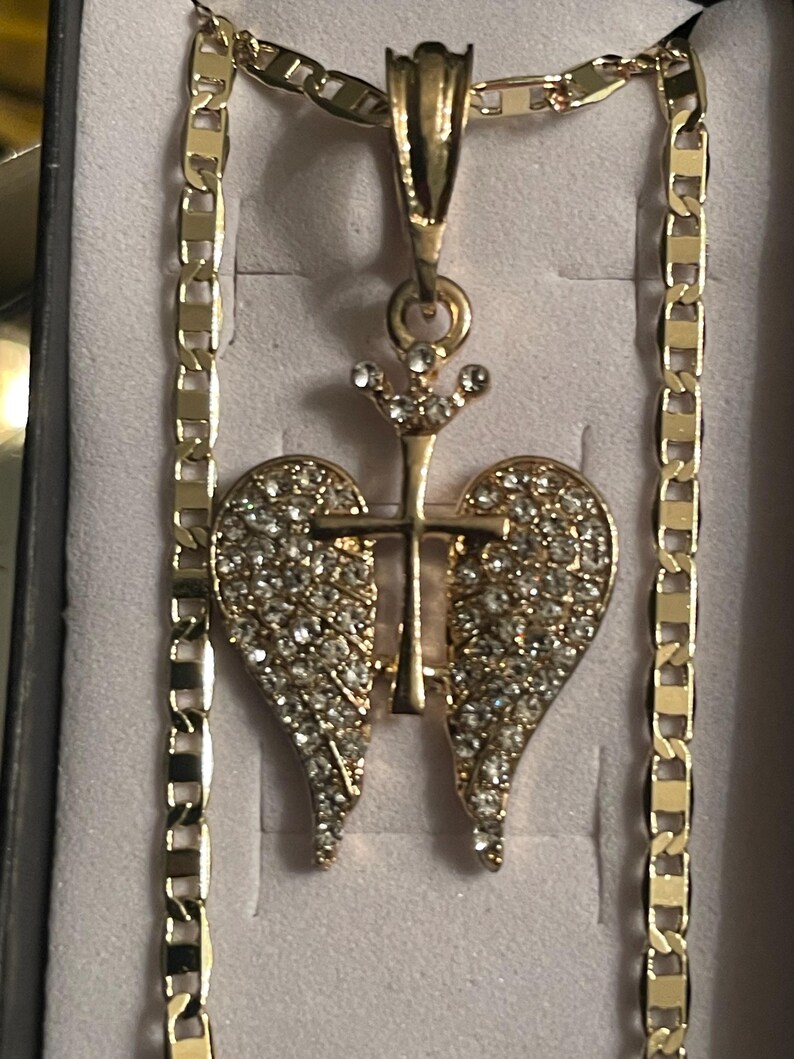 May include: Gold-coloured necklace with a pendant featuring a cross and angel wings design, encrusted with sparkling rhinestones. The chain has a flat, rectangular link pattern. The pendant is centered on a light-coloured background.