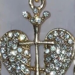 May include: Gold-coloured pendant necklace with a cross design and two heart-shaped wings, both set with sparkling crystals. The pendant has a decorative bail and is likely made of a metal alloy.